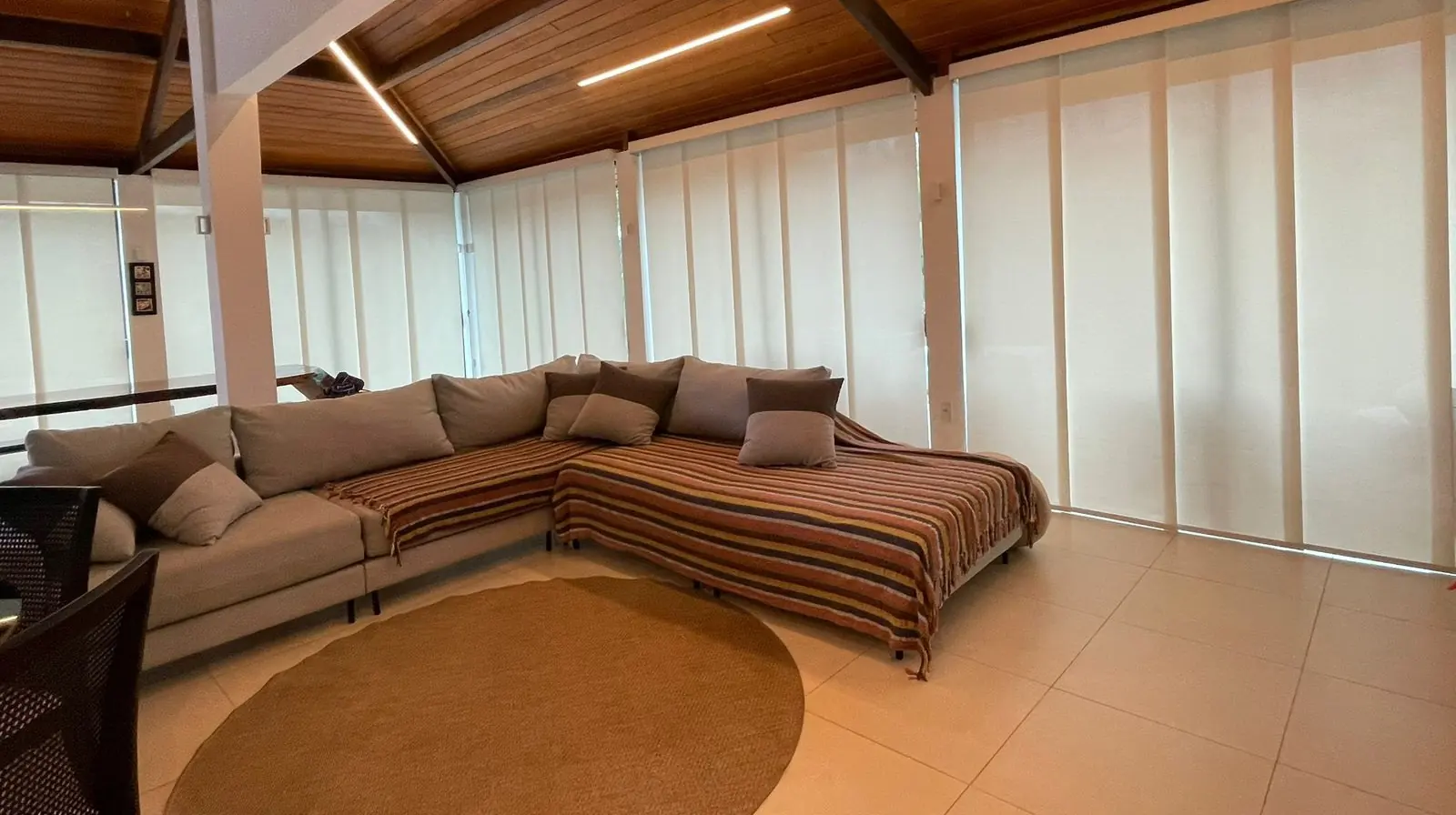 Sala ampla com sofá em L e painéis de tecido claro cobrindo as janelas do chão ao teto